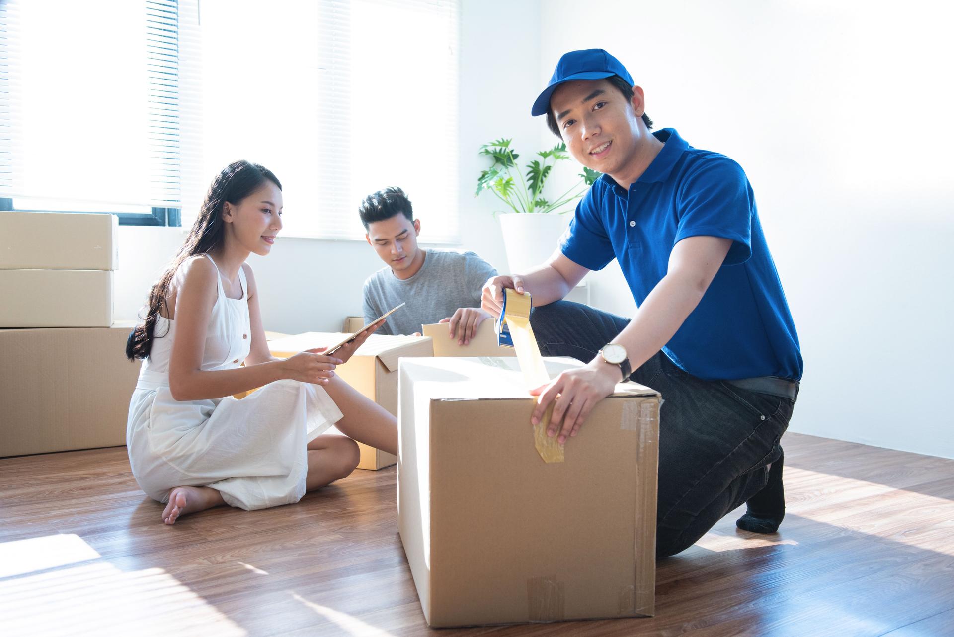 Delivery service worker man in blue uniform help happy couple customer packing parcel with tape in customer home.Woman with tablet signing proof document,man unpacked receive box in background.
